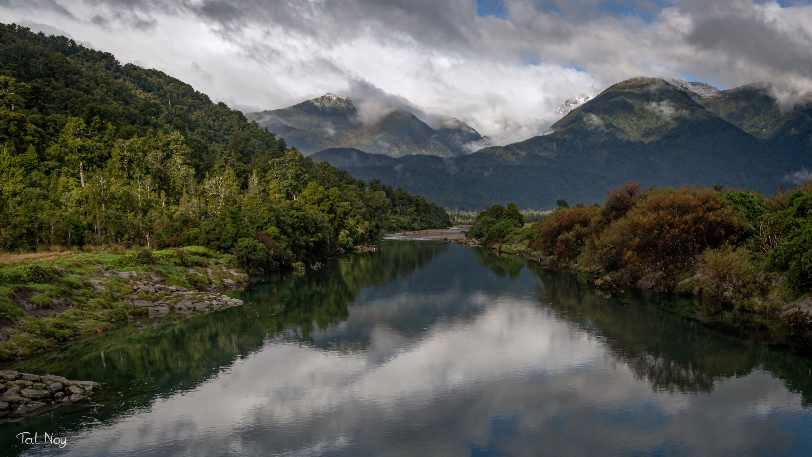 A still river perfectly reflecting clouds and mountains, flanked by lush green forest